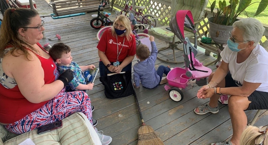 A Save the Children worker along with two other women, sit on a porch with two young kids, surrounded by toys.