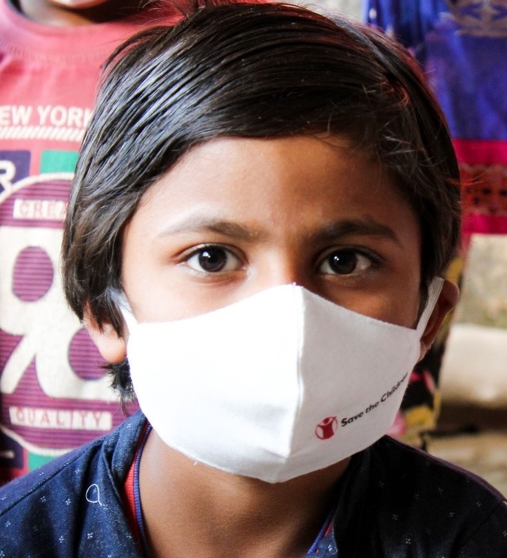 Young boy in a blue shirt looks into the camera wearing a Save the Children face mask. 