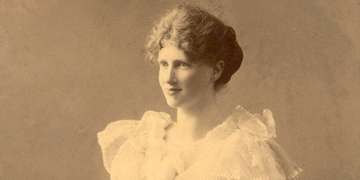 An early, sepia-toned photo of Save the Children’s founder, Eglantyne Jebb. Jebb is dressed in a ruffled, feminine dress, but she kneels on an ornate, wooden armchair – rather than sitting demurely –indicative of her defiant nature. Photo credit: Save the Children.