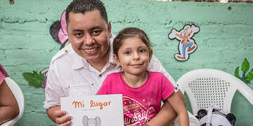 A father and daughter hold a children’s picture book while standing together in front of a green mural decorated with characters.