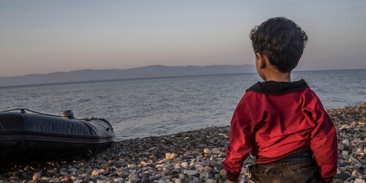 A young boy looks out to sea from the shores of the Greek island of Lesvos. Next to him is an inflatable boat. 