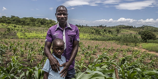 A father stands with his son in a field in Mozambique where he works to support his family