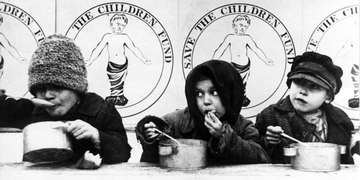 An early black-and-white photo depicts three young children hungrily spooning soup out of tin bowls. The children are bundled up in winter coats and hats; behind them are posters featuring a rudimentary logo for “Save the Children Fund.” Photo credit: Save the Children. 
