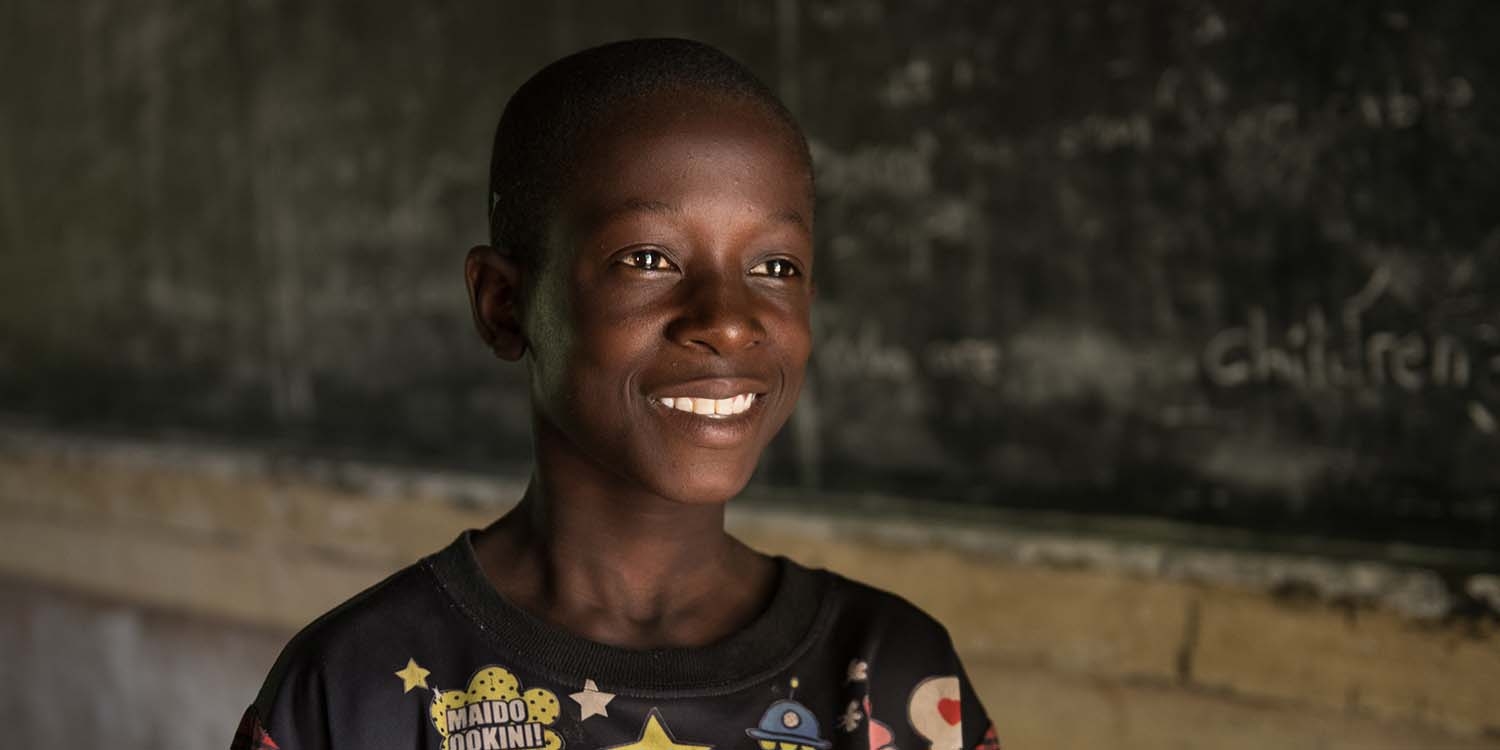 Malawi, a boy in a black t-shirt smiles while in front of a chalk board in a classroom. 