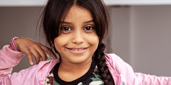 Eight-year-old Johaumi smiles brightly, dressed in a pink sweatshirt and her dark hair in a long braid. Johaumi is a Venezuelan migrant living with her mother and siblings in a dusty border town in northern Colombia. The area is a hotbed of trafficking and brutality and is now home to thousands of Venezuelan migrants who have been driven out of their country by hunger, extreme poverty and violence. They sleep on the street. Photo credit: Sacha Myers / Save the Children, October 2018.  