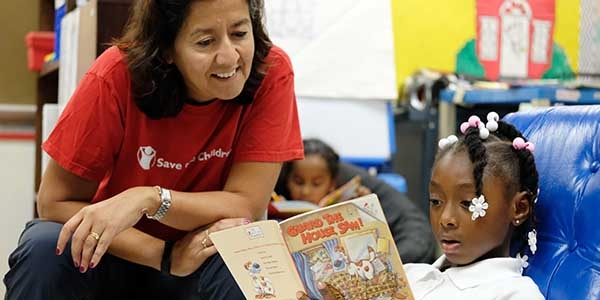 CEO Janti Soeripto wears a Save the Children shirt and looks on as a young girl reads a book. 