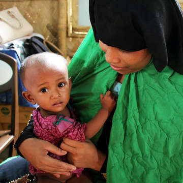 A 1-year-old baby girl clings to her mother at a Save the Children nutrition center in Cox’s Bazar, Bangladesh. Rohingya refugees – the mother is waiting to get a dose of Vitamin A for her daughter as part of Nutrition Action week.  Photo credit: Daphnee Cook / Save the Children, July 2018. 