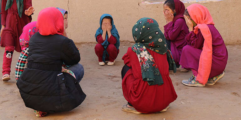 A group of children crouch in a circle, covering their faces with their hands. 