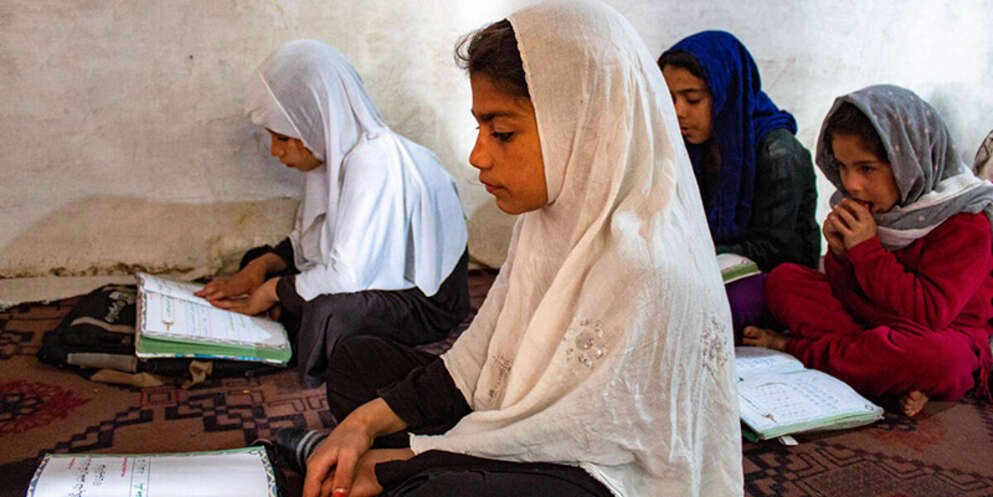 A girl in a white headscarf sits on the floor and reads a book. Her classmates sit around her, also reading.