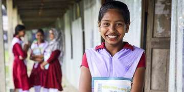 A girl holding a textbook smiles while standing in the courtyard outside her school in Bangladesh.