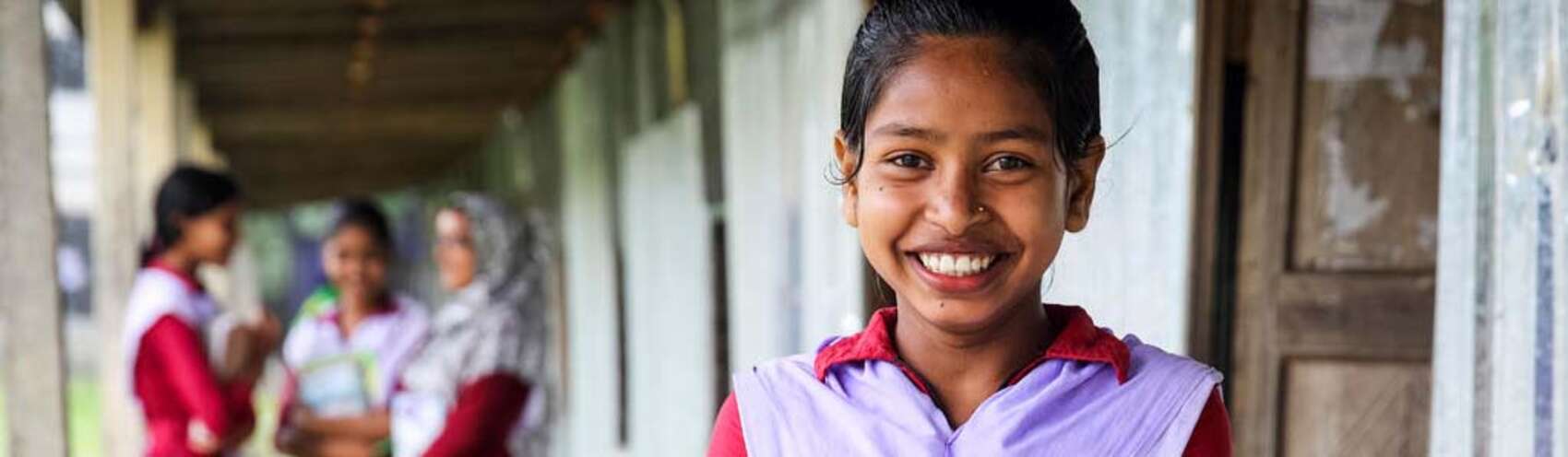 A girl holding a textbook smiles while standing in the courtyard outside her school in Bangladesh.