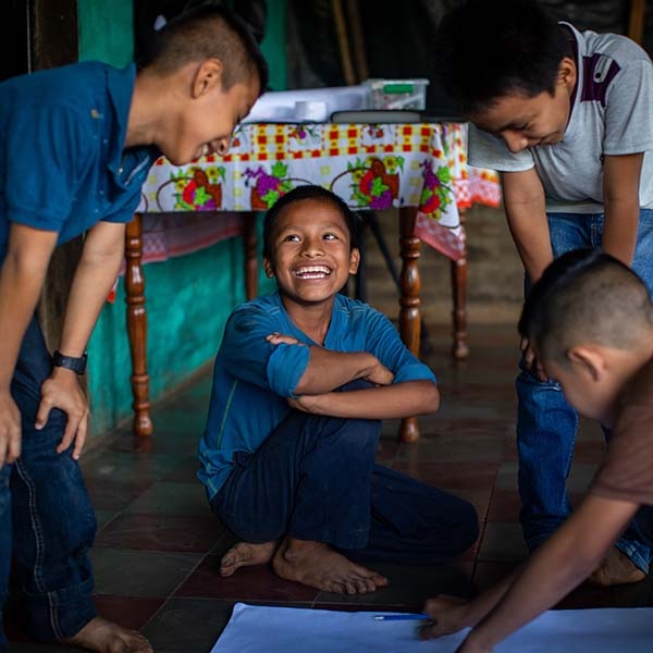 A group of boys in blue shirts laugh together, while doing school work