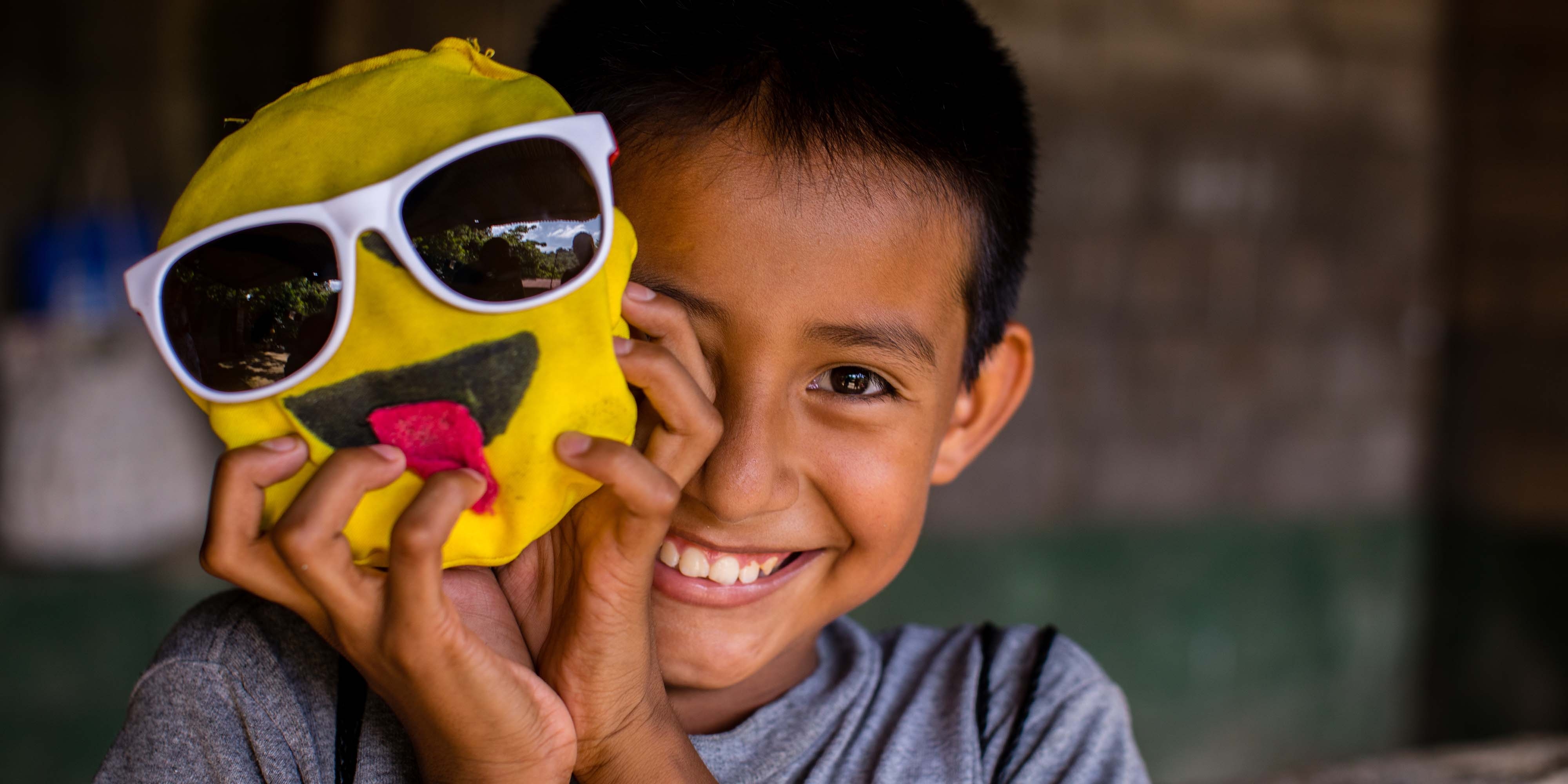 A boy in El Salvador smiles while holding a craft of a yellow smiley face in front of him.
