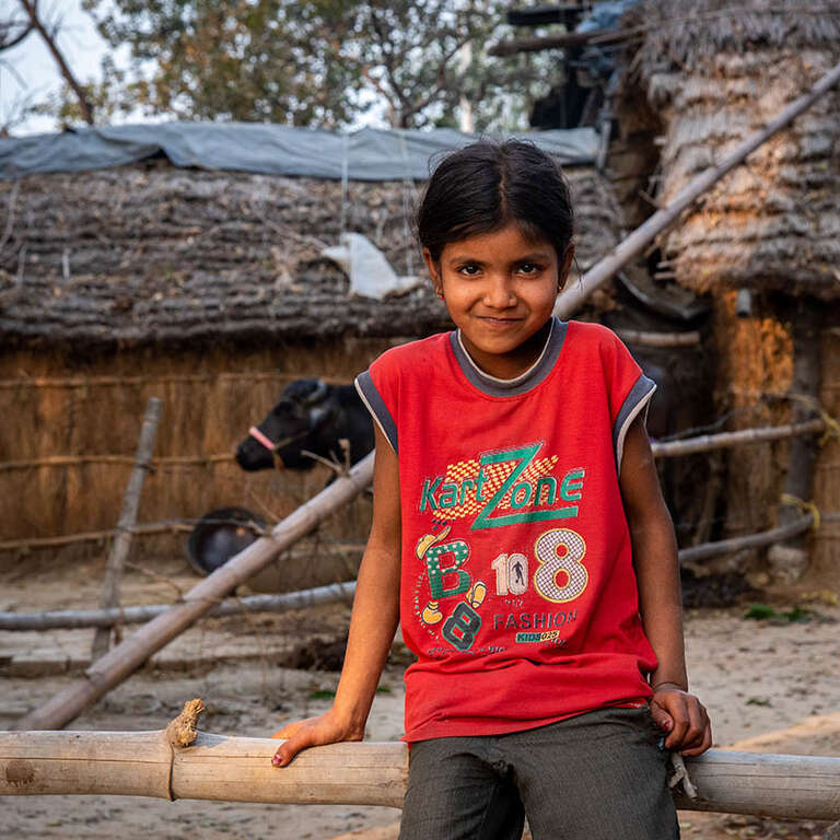 7-year-old Gudiya in a red t-shirt smiles and leans against a wood fence. Her house and a cow are in the background.