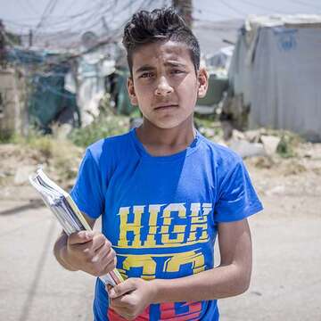 Siraj, age 13, looks concerned, as he stands in the informal, tented settlement where he lives, holding some notebooks. 