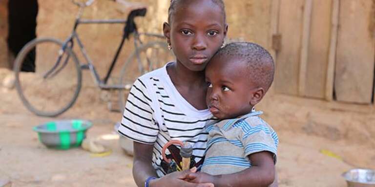 An 8-year old girl holds her baby brother while sitting outside her family's home in Mali.