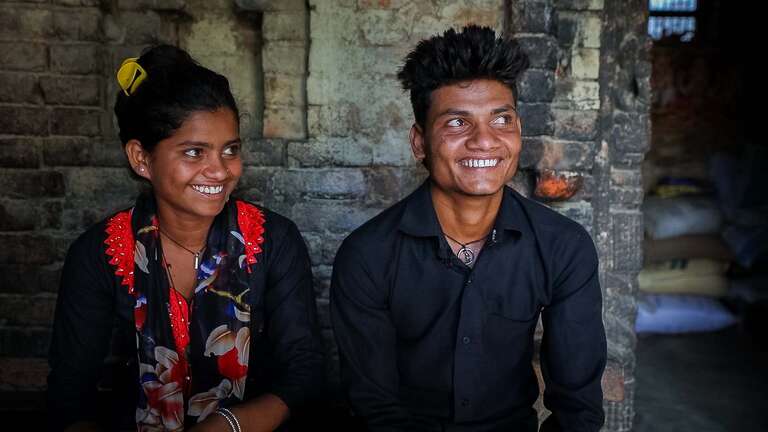 A brother and sister sit next to each other smiling. 