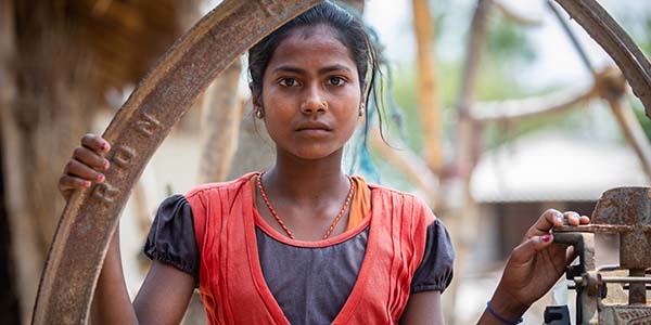 Mamta, age 10, wearing a simple red and blue dress and a serious expression, stands outside her family home in Nepal, worrying about child marriage.