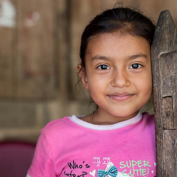 Six-year-old Manela, wearing a pink shirt, smiles as she leans against a wooden door. 