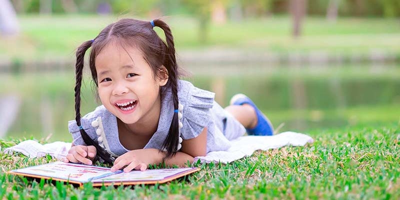 A little girl reads a book in the grass, while smiling at the camera