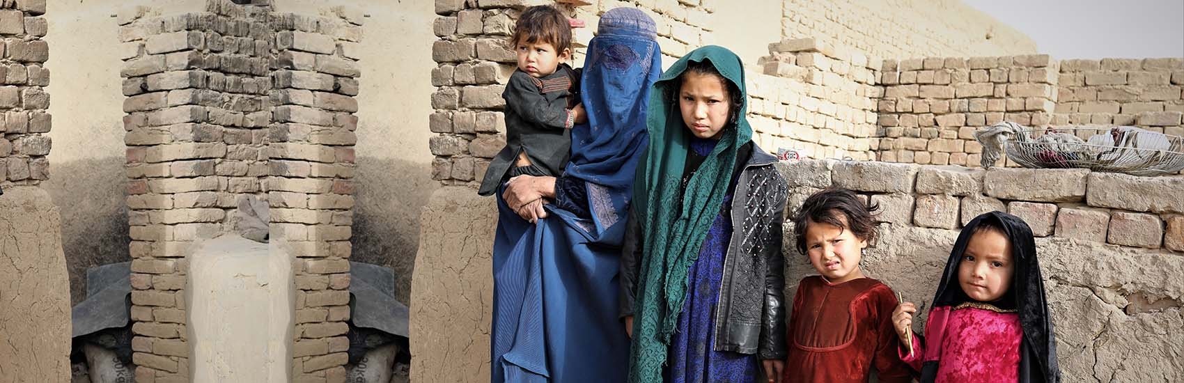In Afghanistan, a mother holds her baby while standing next two her three daughters in front of a crumbling brown brick wall.