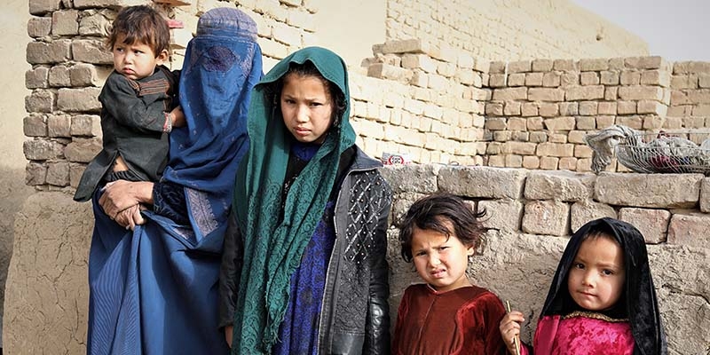 In Afghanistan, a mother holds her baby while standing next to three of her daughters in front of a crumbling brick wall.