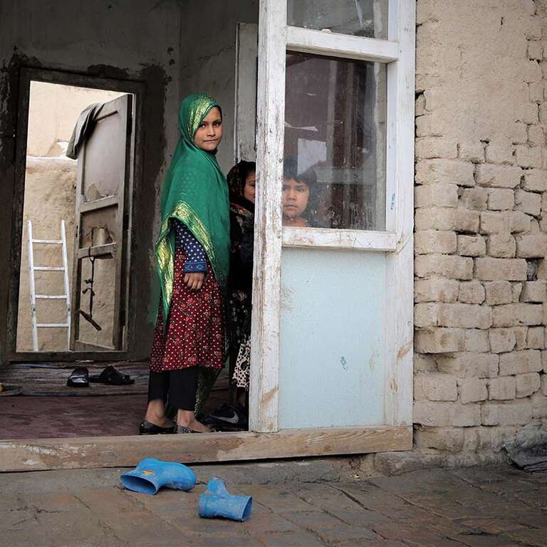 In Afghanistan, a 9-year old girl stands behind an open glass door of a crumbling building while her younger siblings look on. 
