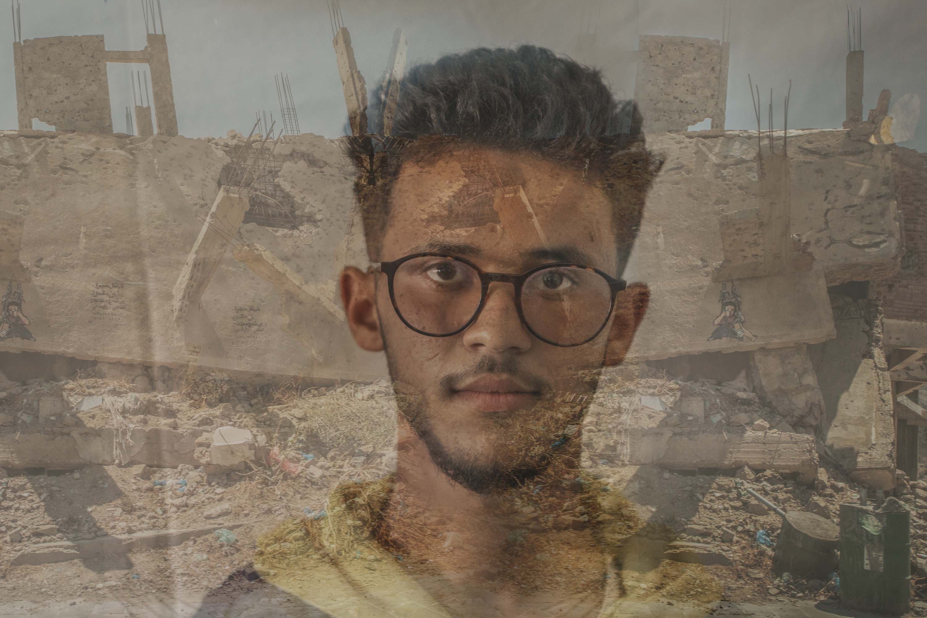 Yemen, a 15 yr. boy in glasses, stands in front of the rubble of a building