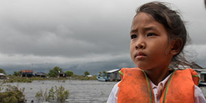A girl in an orange life jacket stands in front of a small village, set on a river. 