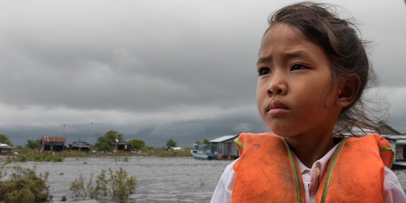 A girl wears an orange life preserver and sits in a boat looking out over the flooded waters. 