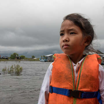 A girl in an orange life jacket stands in front of a village, set on a river. 