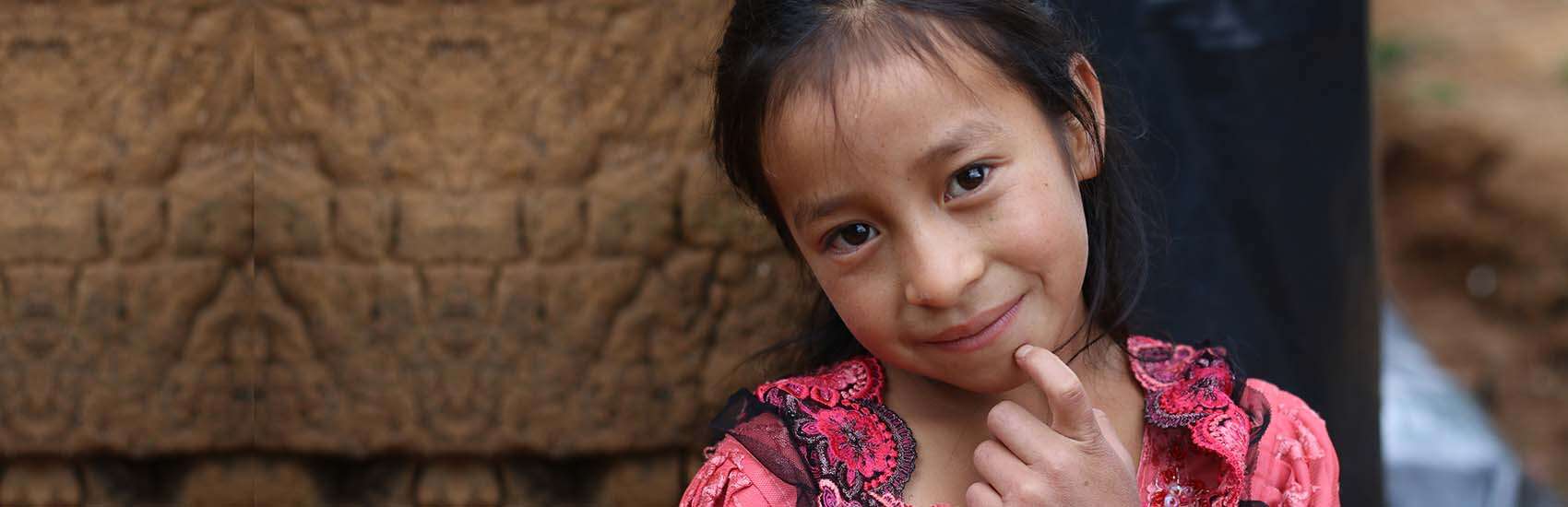 In Guatemala, a girl smiles while holding a finger up to her face.