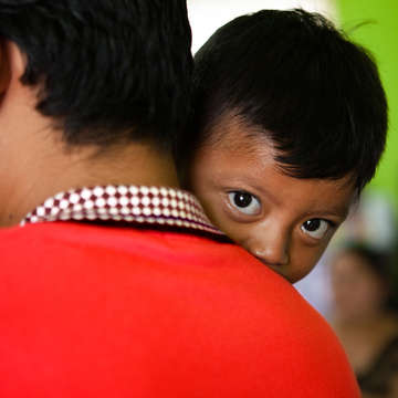 A young child from Guatemala rests his chin on the shoulder his father. The child was displaced from his home following a volcanic erruption. Many families and children from Central America, including Guatemala, have been forced to flee their homes due to conflicto, disaster and poverty. Some families have chosen to migrate to the United States in search of a better life, however the crisis at the U.S.-Mexico border is causing families to be seperated, children to be detained for long periods of time, and more. Photo Credit: Save the Children Guatemala