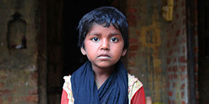 A young girl in a red shirt and blue scarf looks into the camera. 