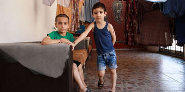 Two boys pose in an empty room. One sits on a couch, the other leans against it. 