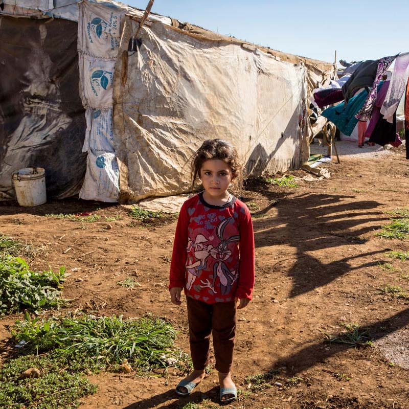 A young Syrian refugee girl stands alone in front of a laundry line and home in a refugee camp.