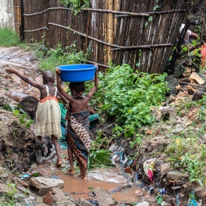 Two young girls, both barefoot and one balancing a large blue basin on her tiny head, step across sharp rocks and muddy water. Next to them is a flattened shelter destroyed by Cyclone Kenneth which struck northern Mozambique on April 25, 2019. Save the Children is scaling up response efforts to provide emergency relief to families and children who have been impacted by this devastating cyclone. Photo credit: Sacha Myers/Save the Children, April 2019. 