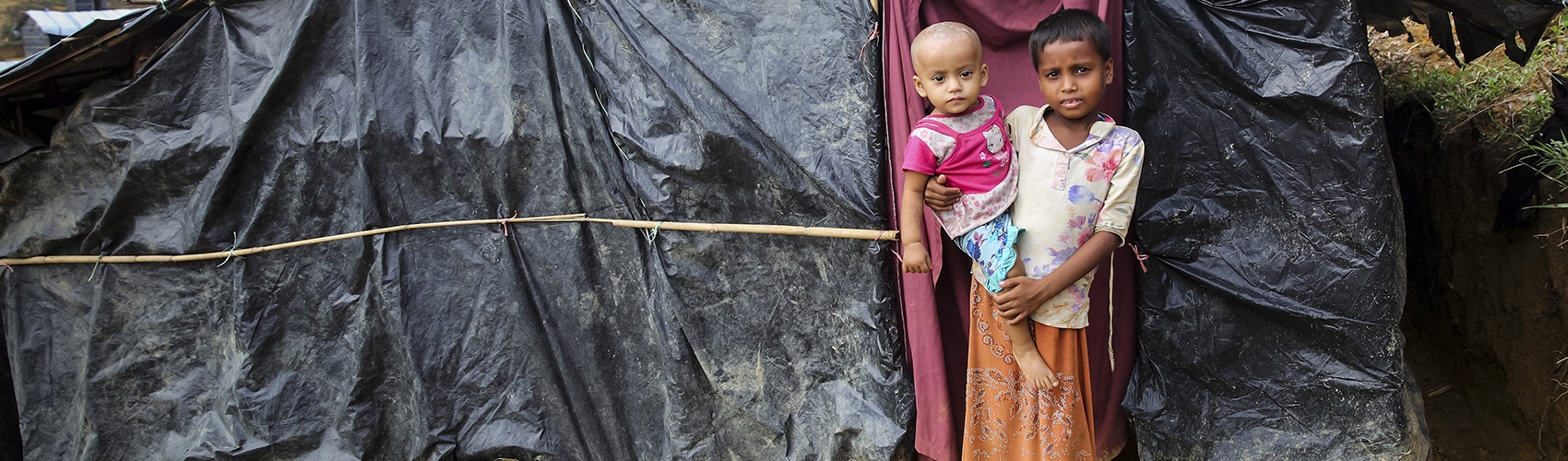 A six-year old boy holds his one-year old brother as they stand outside the makeshift tent in Cox's Bazar district, Bangladesh where they live.