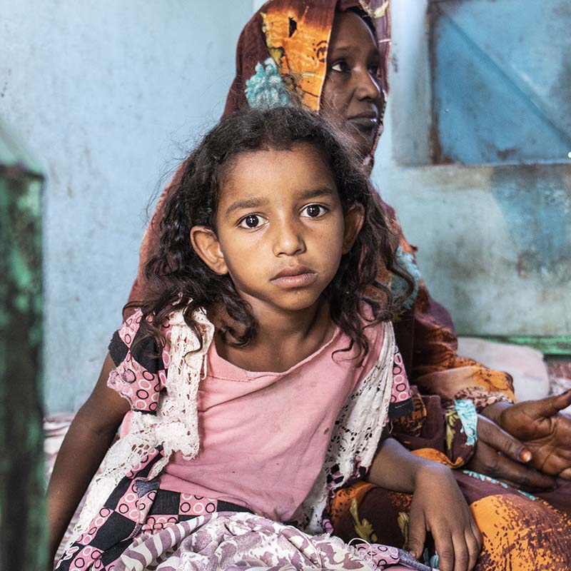 In Sudan, an 8-year old girls sits with her mother in a health facility. 
