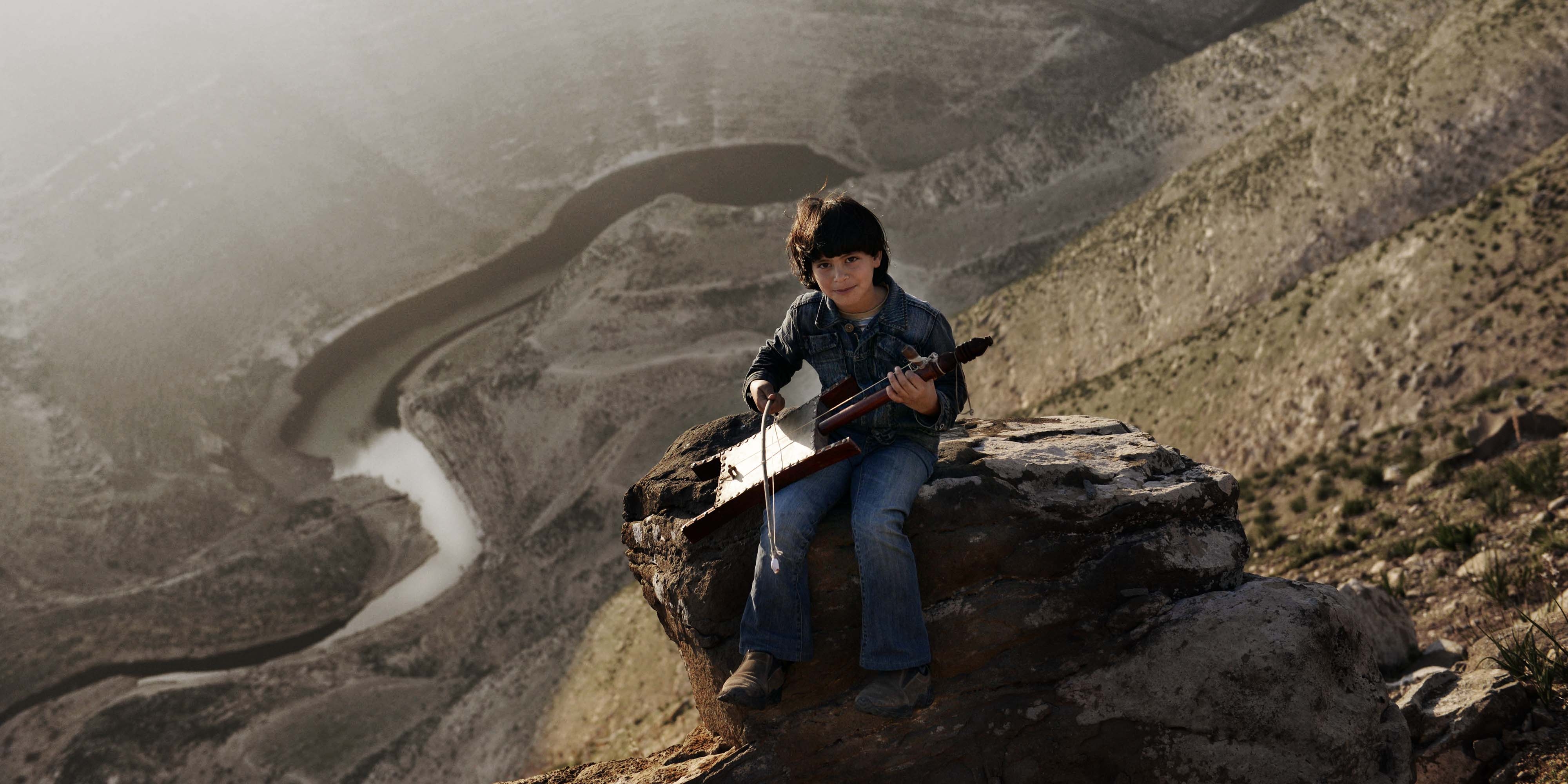 Syria, a boy in jeans sits on a rock on a hillside holds an instrument