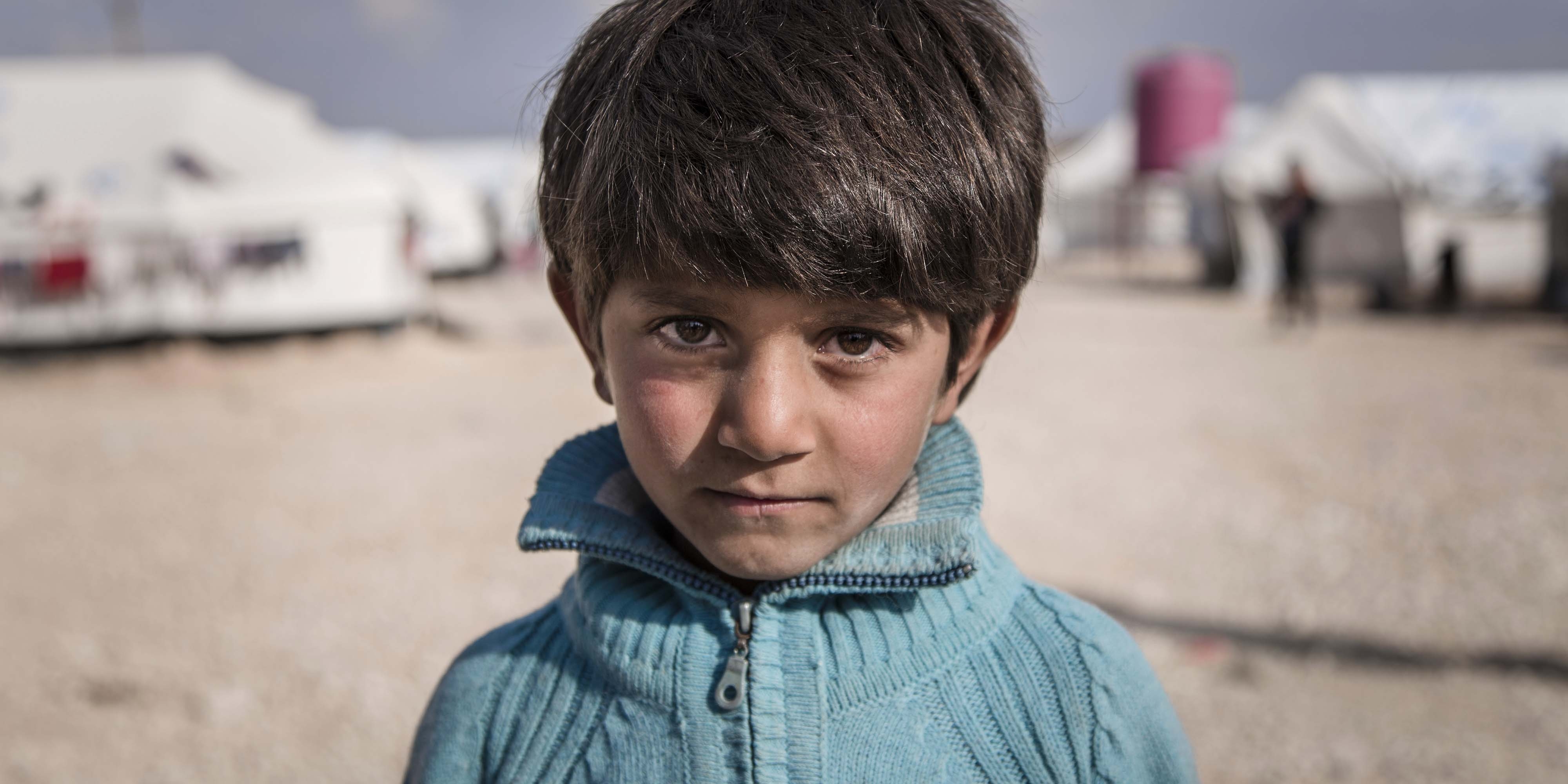 Syria, a little boy in a blue sweater stands in front of his family's tent in the Al Hol camp