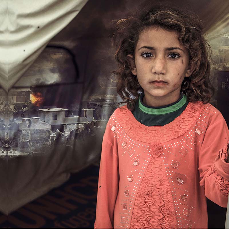 A Syrian girl stands in front of a makeshift shelter as a scene of fire and destruction is shown in the background.