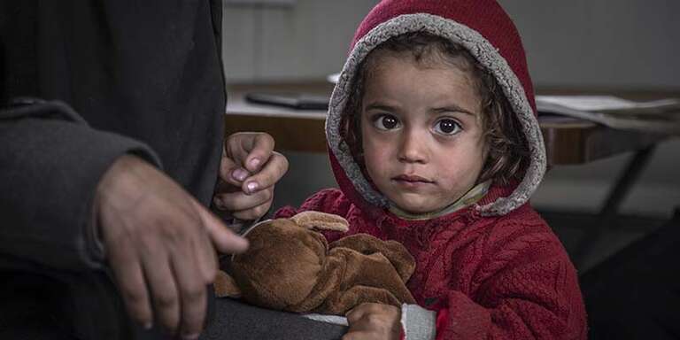 In Syria, a young girl displaced by conflict sits with a stuffed animal and wears a hooded sweater to stay warm. 