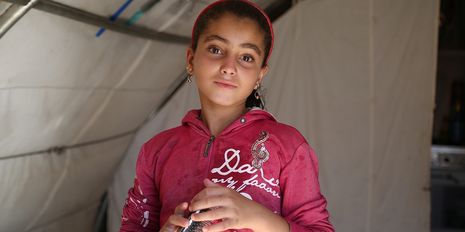 In Syria, a girl stands in a tent after being displaced from her home due to conflict. 
