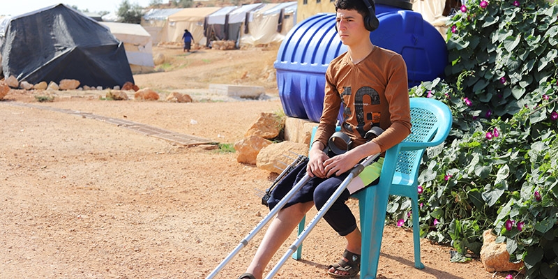 Syria, a teen boy sits  outside his tent, listening to music.