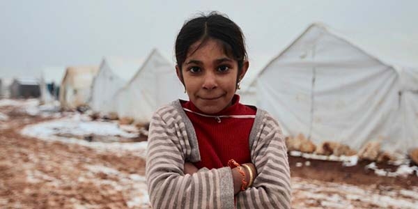 A young girl stands with her arms folded. Behind her, snow covers the ground and a row of white tents can be seen in the background.