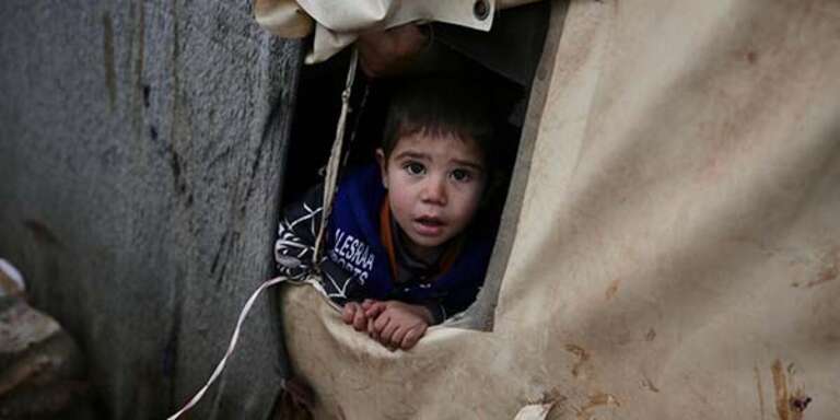 A young boy without a coat peeks his head out of a makeshift shelter, erected in an overcrowded camp in Idlib, Syria. 