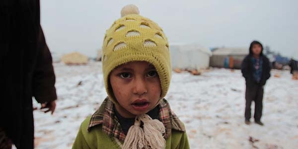 A young boy in a yellow hat standing in a snow-covered displacement camp in North West Syria.
