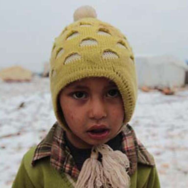 A boy in a yellow hat and green sweater stands in a snow-covered displacement camp in North West Syria. 