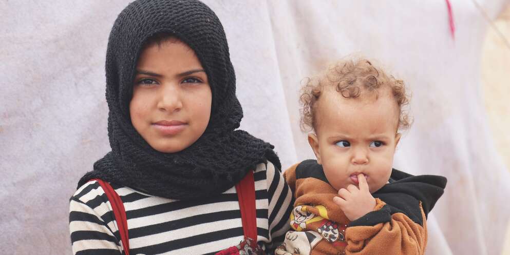 A young girls holds her sibling on her hip while standing outside a tent in a refugee camp in Syria